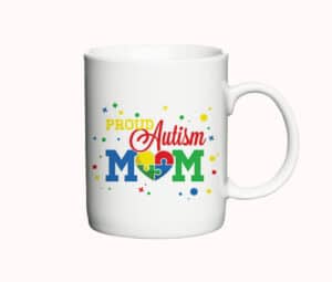 Hvid kaffekop med teksten "Proud Autism Mom" i farverig typografi og et puslespilshjerte i centrum, omgivet af små farvede mønstre.