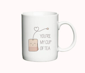 Hvidt keramisk krus med en illustration af en smilende tepose, en hjerteformet snor, og teksten "You're My Cup of Tea".