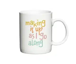 Hvid krus med teksten "Making it up as I go along" i farverige bogstaver.