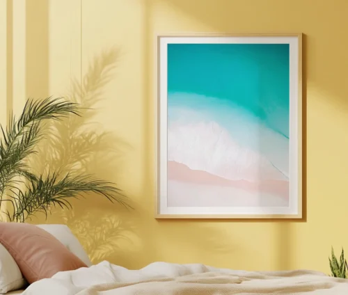 Plakat med strandmotiv hængende på en gul væg i et soveværelse med sollys.