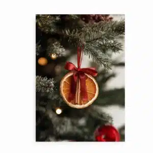 Plakat med tørret appelsinskive og kanelstang bundet med rødt bånd på juletræ.