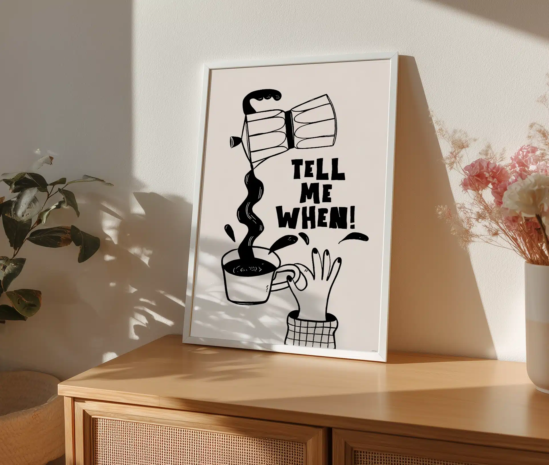 Sort-hvid kaffeplakat stående i ramme på skænk omgivet af planter og blomster.