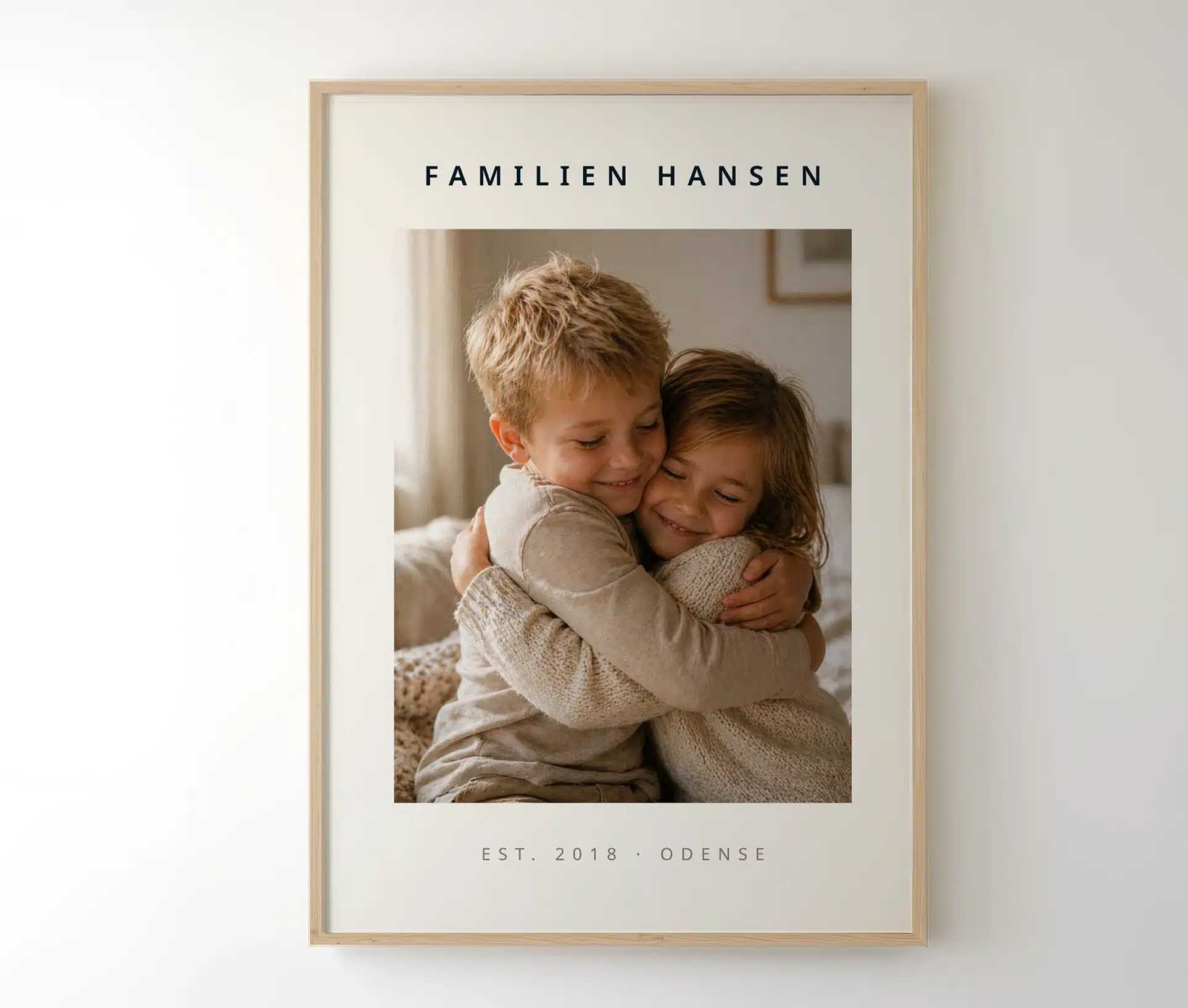 Familieplakat i lys træramme med søskende-foto og familienavn hængende på hvid væg